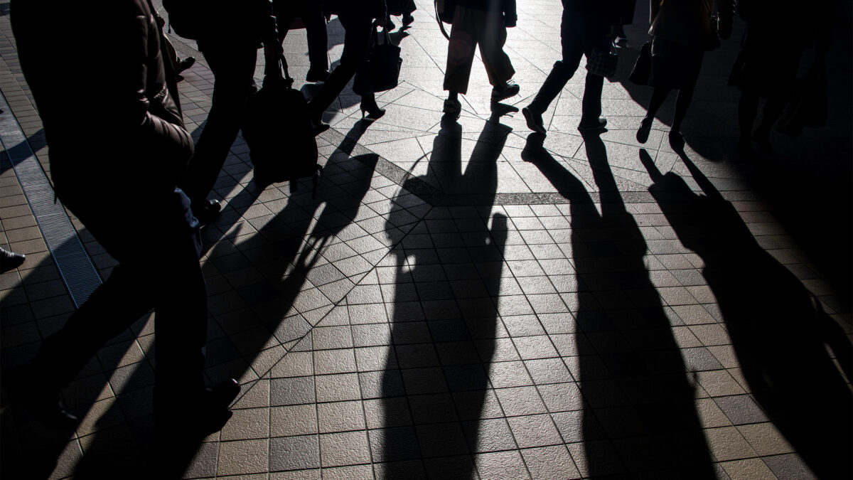 A photo of silhouettes of commuters heading to work from a train station. The morning light makes them cast long shadows.