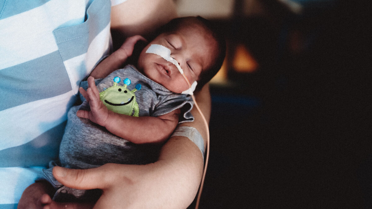 A premature newborn is cared for in the neonatal intensive care unit while being held in a person's arms.