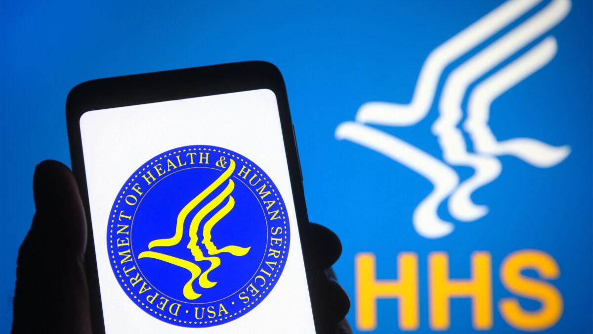 A photo of someone holding up an image on their phone of the Department of Health and Human Services logo. Behind them is a backdrop with the same logo.