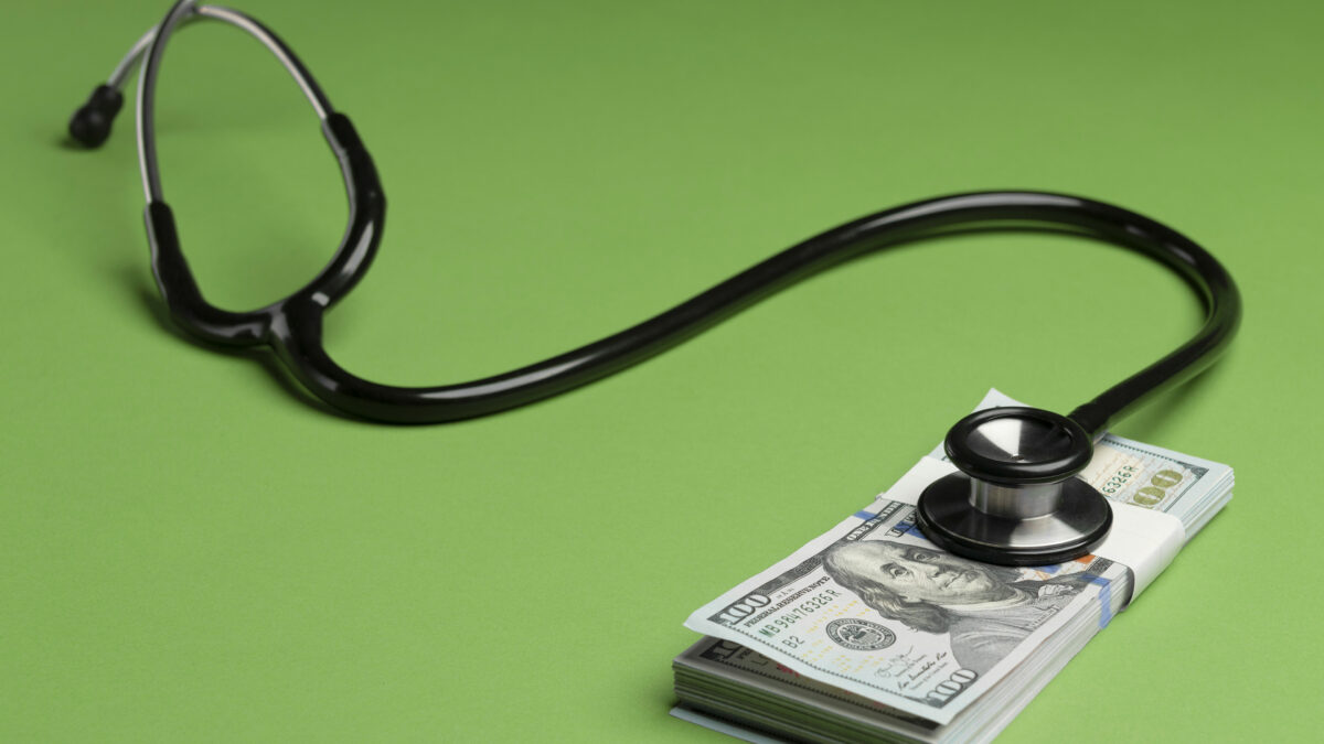 A photo of a stethoscope against a green background. It is resting on top of a stack of $100 bills.
