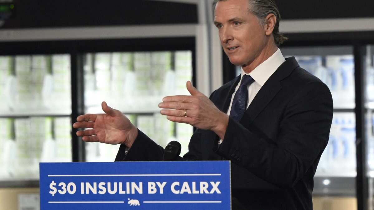 California governor Gavin Newsom stands behind a blue and white sign that reads "$30 insulin by CalRx"