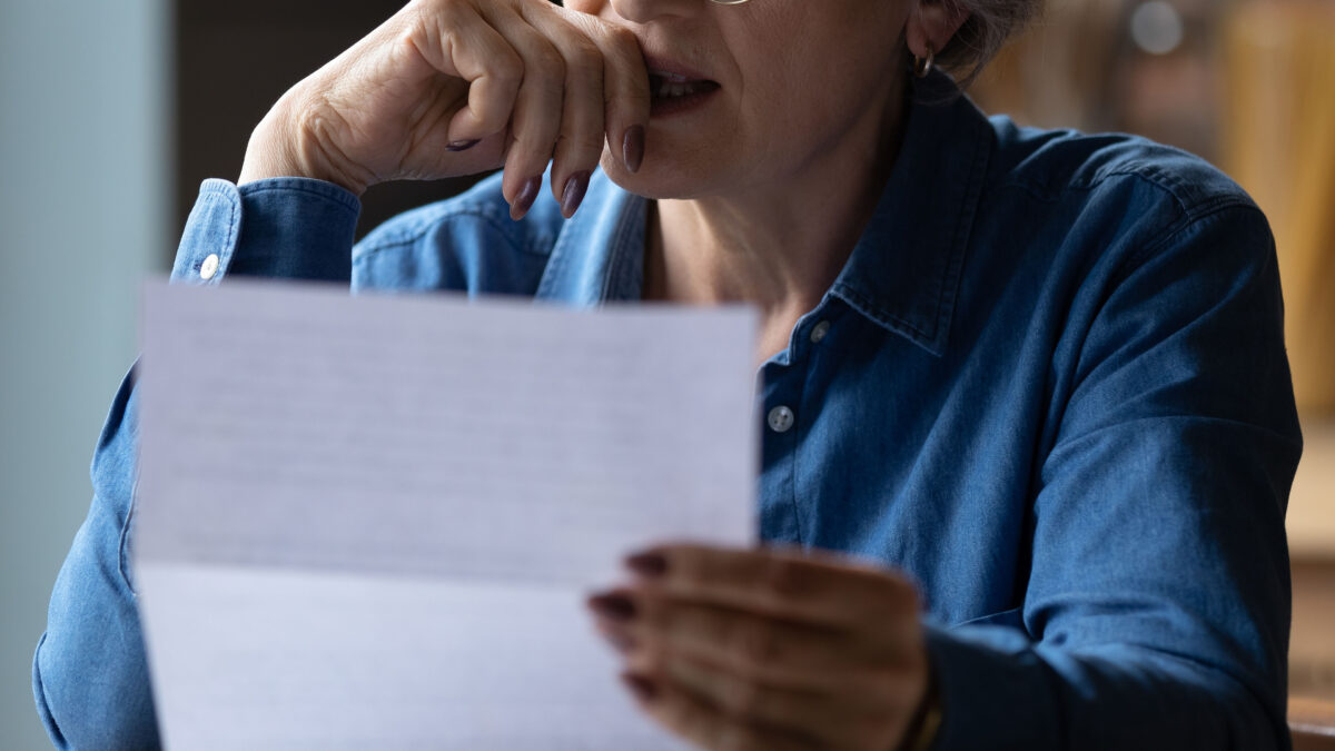 A closely cropped photo of a senior woman holding a paper letter. She presses her hand to her lips as she makes a stressed expression.