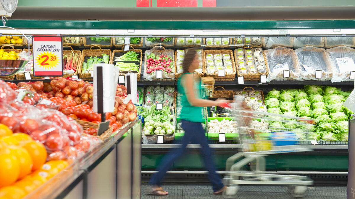 A photo of a woman pushing a grocery cart through a produce aisle in a grocery store.