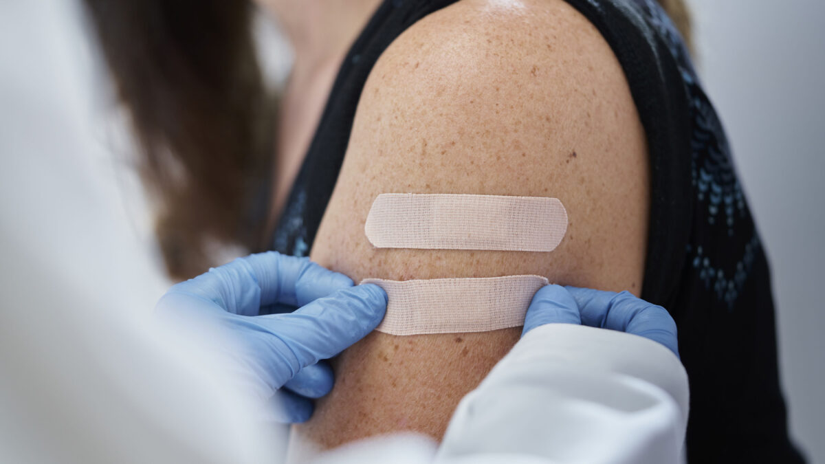 A close-cropped shot of a doctor wearing blue gloves giving second adhesive bandage to a young woman after administering vaccines.