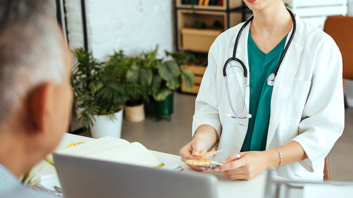 A young female doctor sits at a desk in an upscale office. A male patient sits across from her. There are plants and modern furniture in the background.