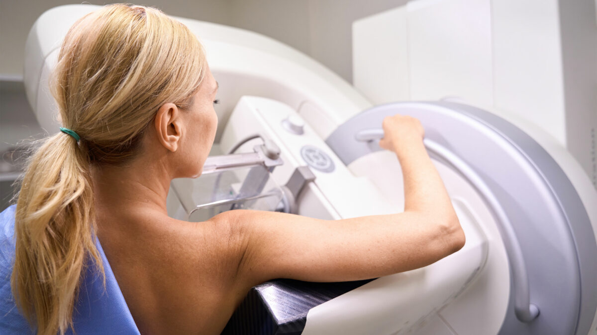 A woman with straight blonde hair stands at a mammogram machine. Her light blue hospital gown is off her right shoulder.