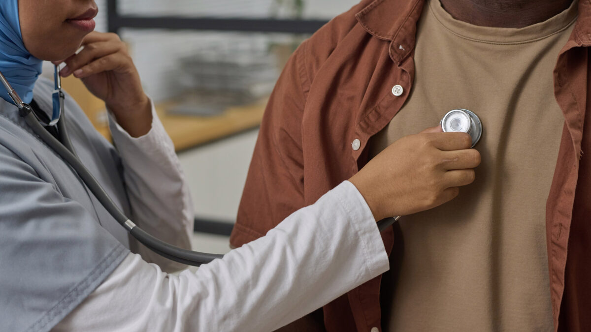 A female primary care doctor examines a male patient using a stethoscope.