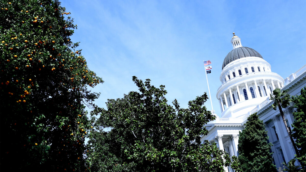 A photo of the California State Capitol at an angle on a sunny day. Orange and palm trees are seen in the foreground in front of the building.