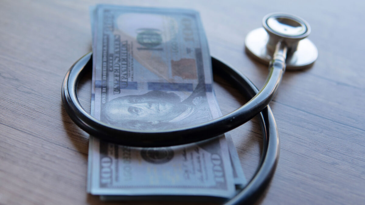 A close-up image of a black stethoscope wrapped around a stack of U.S. dollar bills.