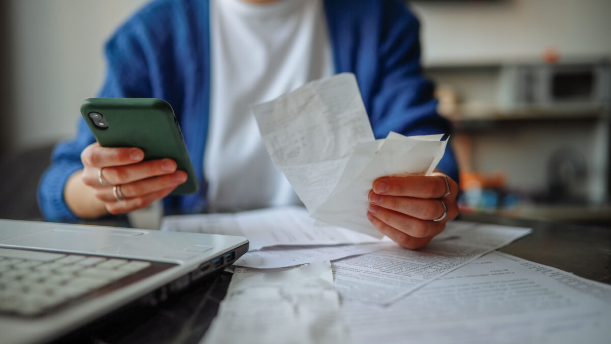 A photo of a person holding receipts and a cell phone while sitting at a table covered in letters and bills.