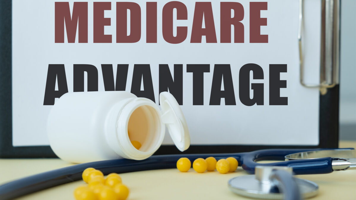 A photo of a clipboard resting horizontally on a table with a piece of paper that reads, "Medicare Advantage." In front of it are an open pill bottle spilled yellow capsules, a stethoscope, and a pen.