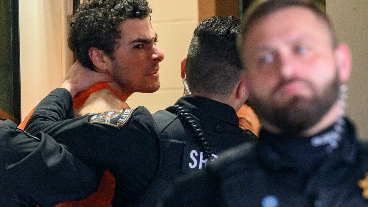 A photo of a man scowling at the camera as he is escorted indoors.