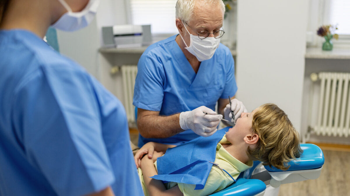 An older male dentist performs a cleaning on a young boy patient. A female hygienist is seen blurred in the foreground.