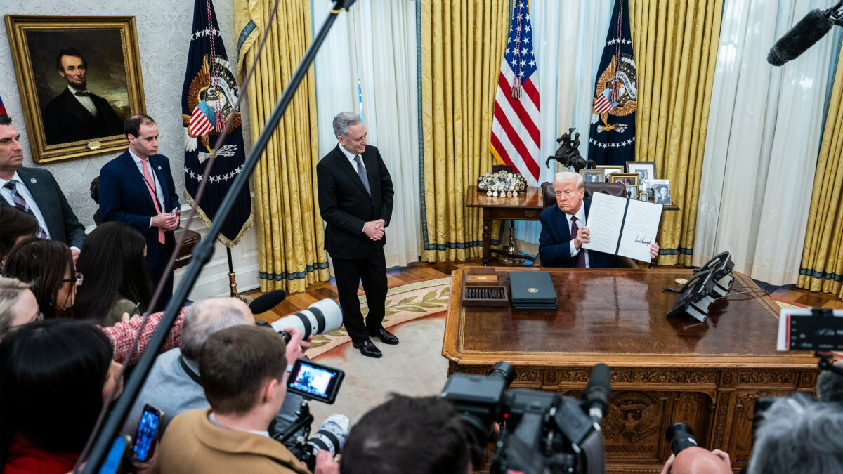 A photo of President Trump holding up a signed executive order while surrounded by news cameras and microphones.