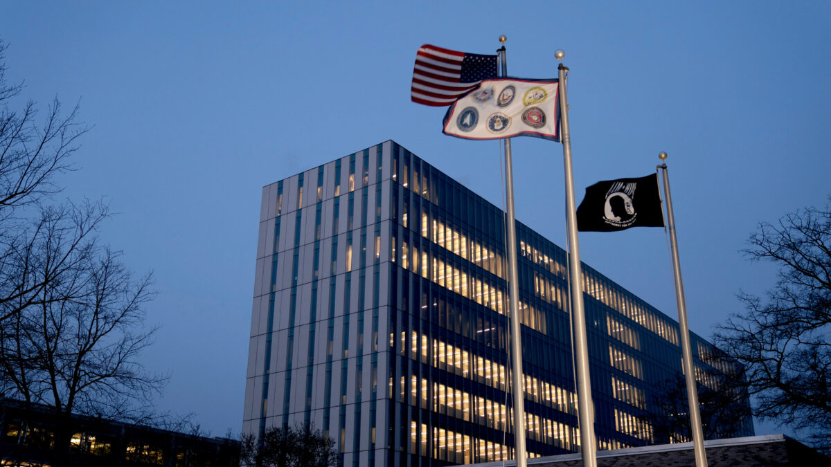 A photo of a large, modern office building in Maryland. In front of it are the American flag, a flag with insignias for six military branches, and the National League of Families POW/MIA flag.