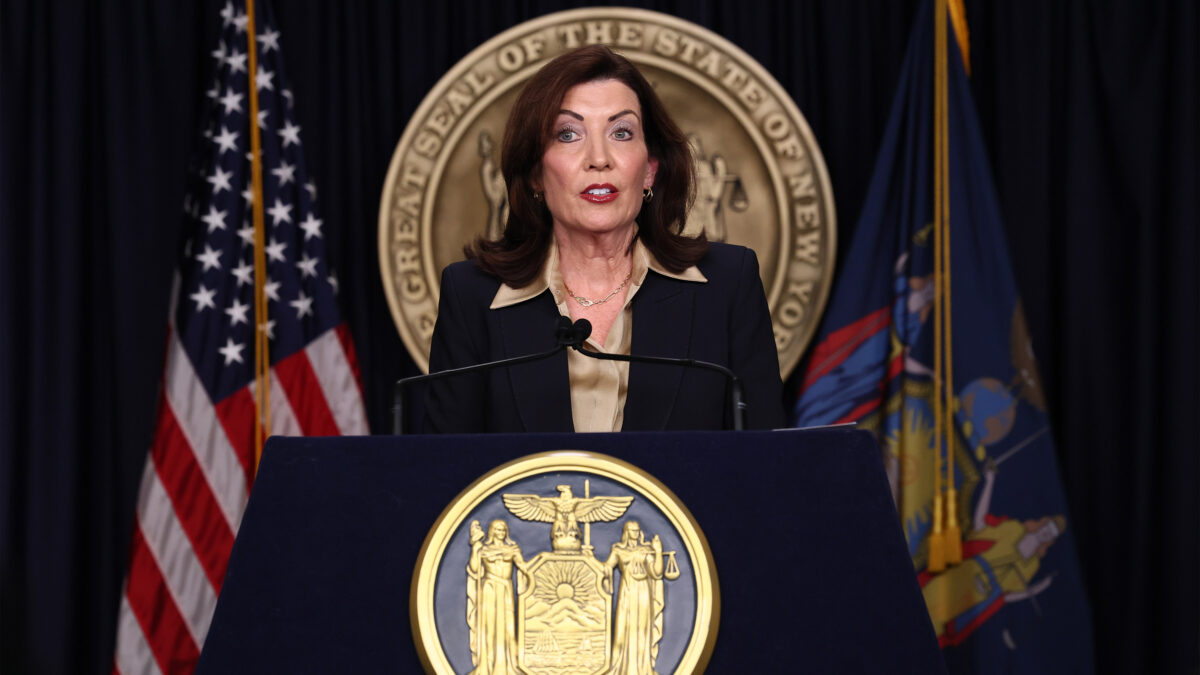 A photo of Kathy Hochul speaking at a podium with the USA and New York flags behind her.