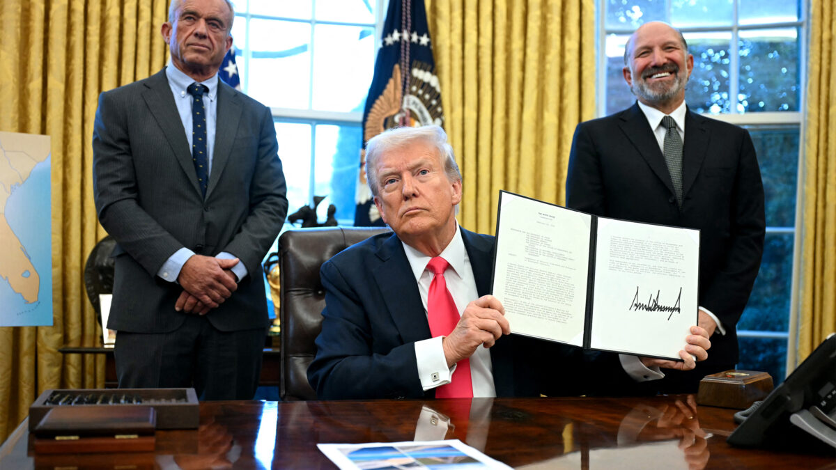 A photo of President Trump showing a signed executive order to photographers in the Oval Office of the White House. He is flanked by Robert F. Kennedy Jr. and Howard Lutnick.