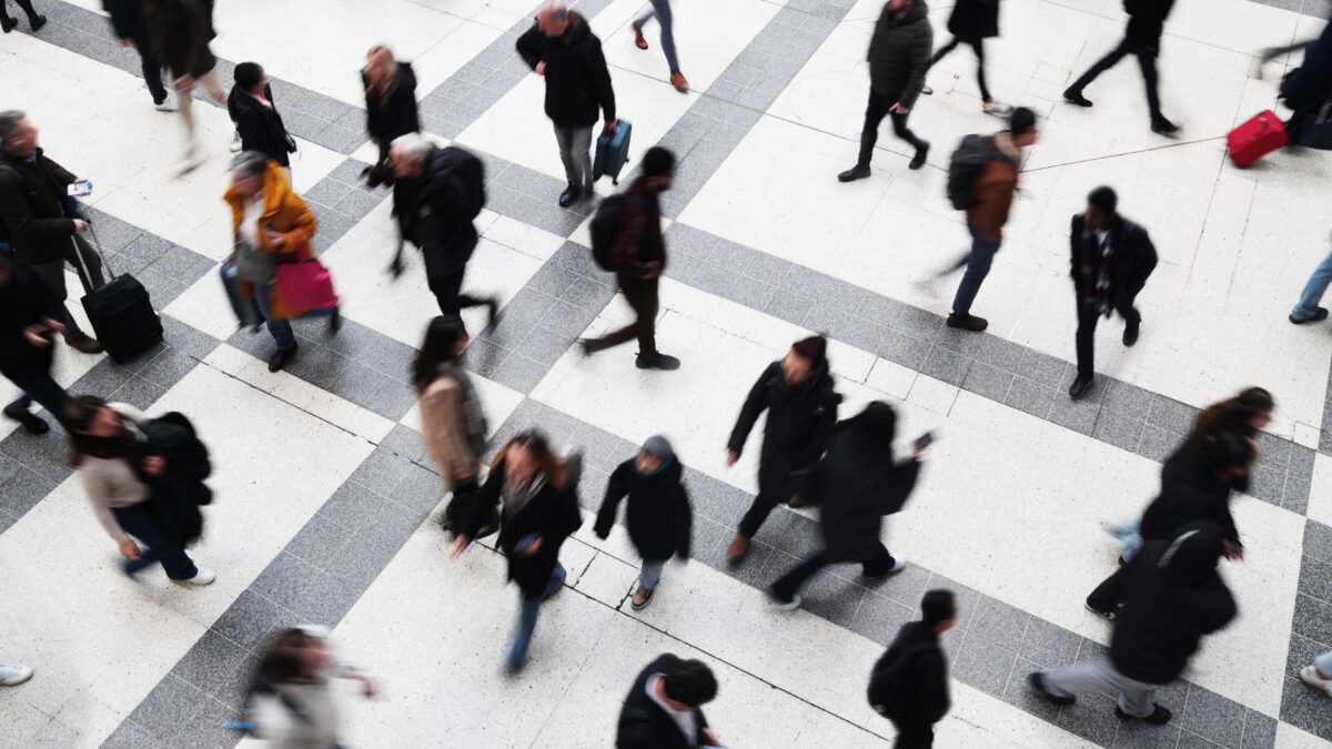 A photo of a train station as commuters and pedestrians walk hurriedly around the frame. Their forms are blurred as they are in motion.