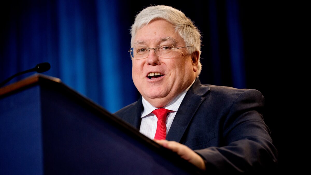 A man with grey hair, glasses, a red tie and a dark suit stands behind a podium