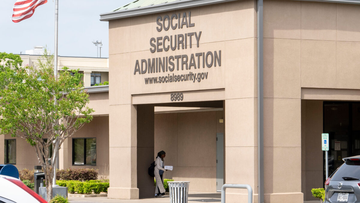 A photo of a concrete office building with the words "Social Security Administration" and "www.socialsecurity.gov" on a sign on the front entrance. An American flag on a pole is posted near the entrance.
