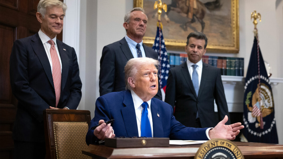 A photo of President Trump seated at a desk, gesturing with his hands, holding a magic marker. Three men stand behind him; they are heads of federal health agencies.