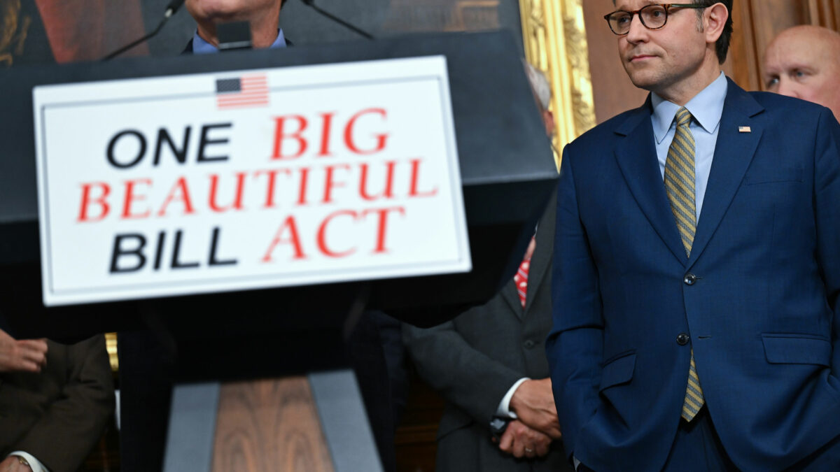 A close-up photo of a lecture with a sign reading "One Big Beautiful Bill Act" on its front. A person stands behind the lecture, but the top half of his face is out of the shot. Speaker of the House Mike Johnson is seen standing to the right of the lecture.