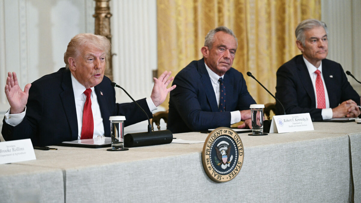 A photo of a White House event: President Trump sits at a table. To the right of him is Robert F. Kennedy Jr. Mehmet Oz sits to Kennedy's right. All three men have microphones across from them.