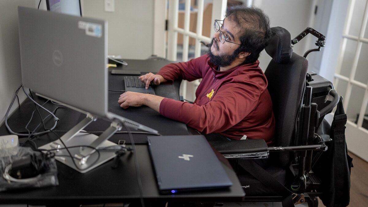A man with muscular dystrophy works at a desk with multiple computer monitors.