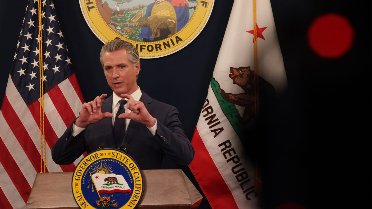Gavin Newsom speaks behind a podium at a press conference. The American flag and California flag are behind him.