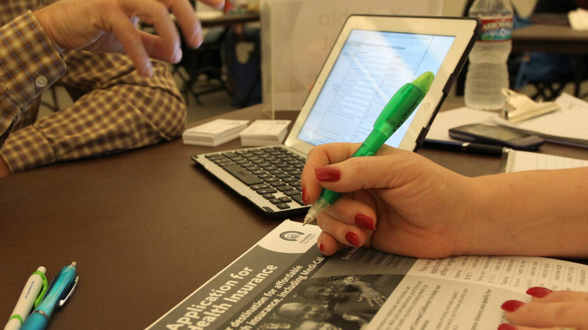 A photo of a laptop on a table. In the foreground is a woman holding a pen over a Covered California information packet.