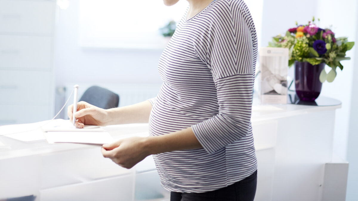 A pregnant woman is at reception for doctor's office, signing a piece of paper.