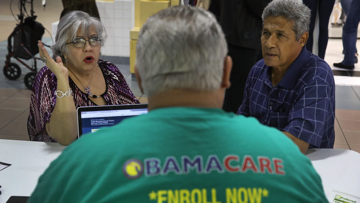 A photo of an older couple seated at a table. A man in the foreground is seen from behind. His shirt reads, "Obamacare. Enroll now."
