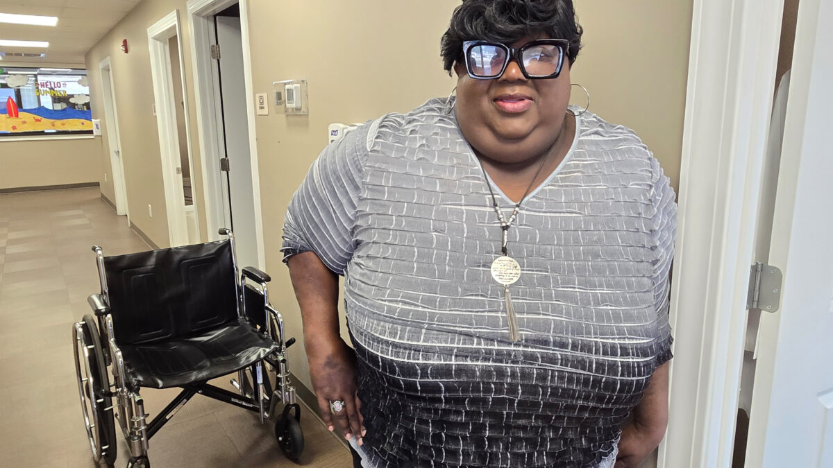 A photo of a woman standing for a portrait in a hallway. A wheelchair is behind her.