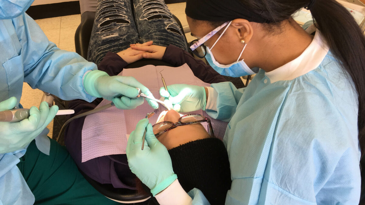 A photo from above of a person lying in a dental chair having work done. A dentist and assistant sit on either side of the patient's head.