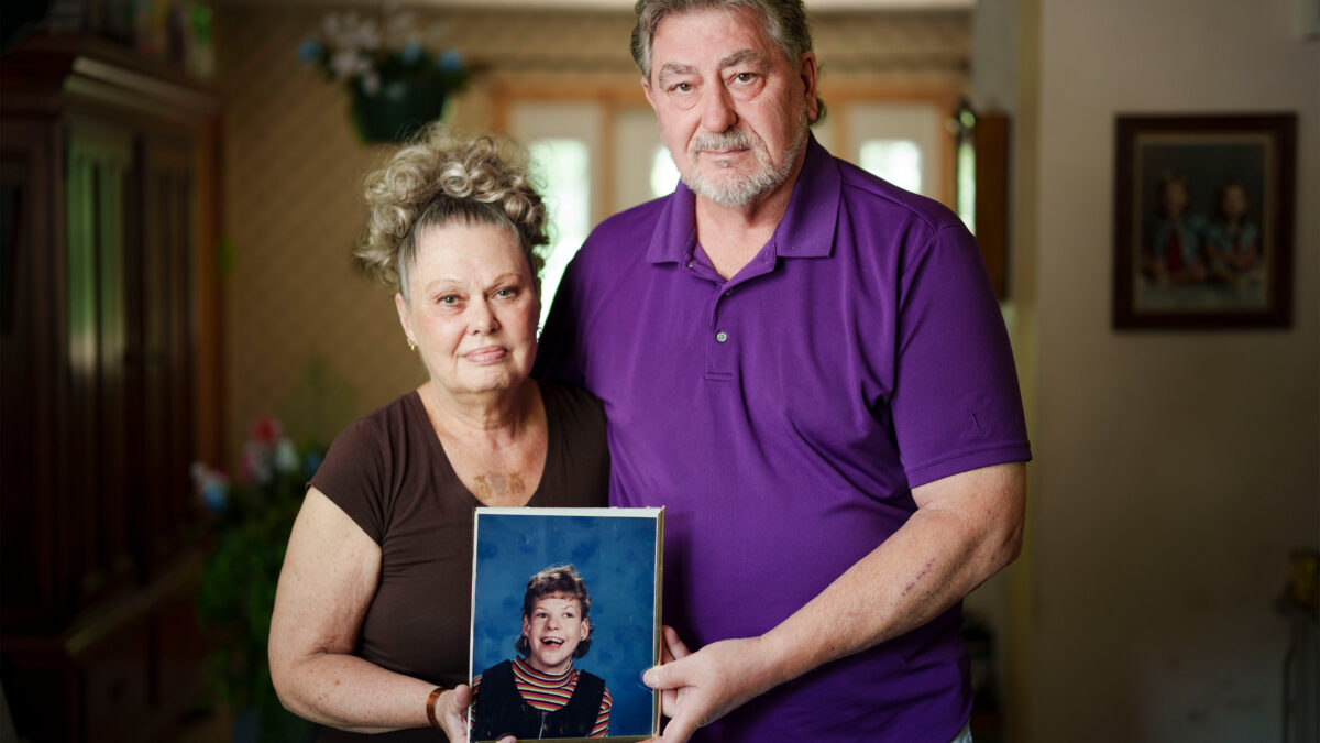 A photo of an older man and older woman standing for a photo in their living room. They're holding a school photo of their daughter.