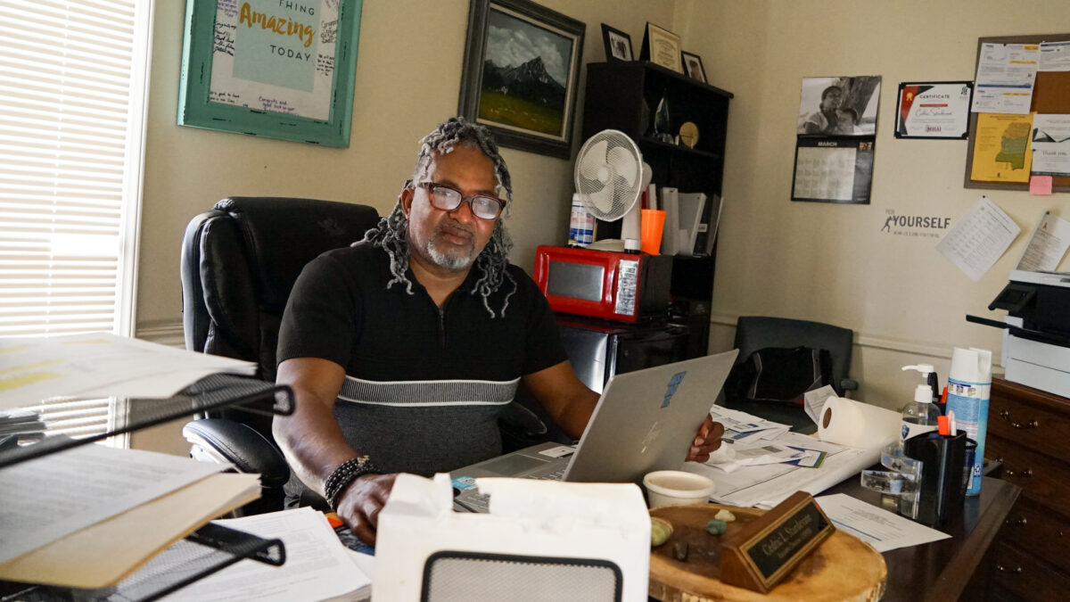 A man sits at an office desk that shows lots of signs of activity; stacks of paper, an open computer, and a name plate.