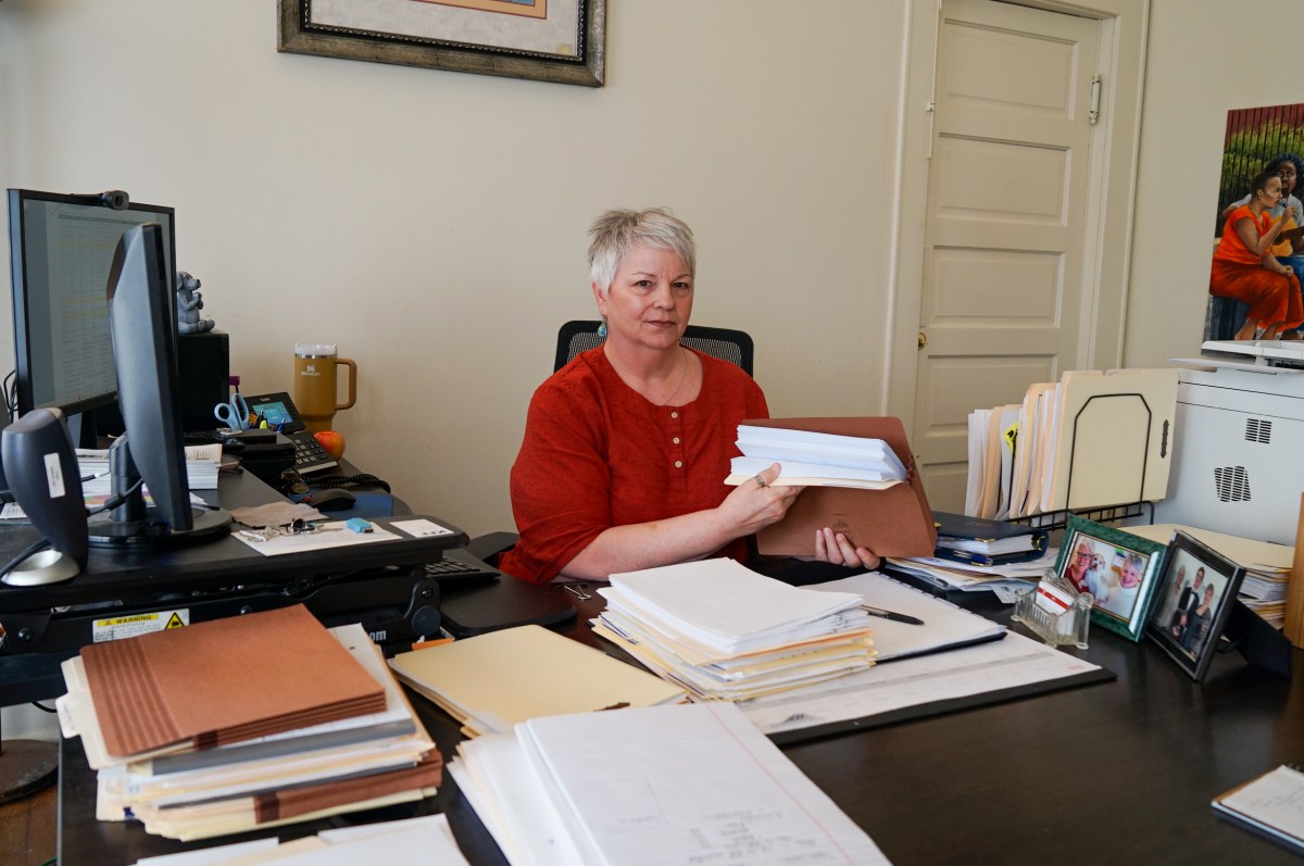 A woman sits at a desk covered in stacks of paper and two computer monitors.