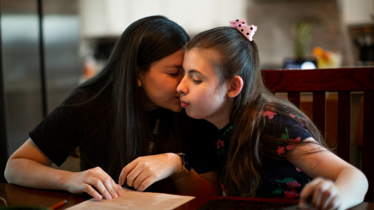 A woman with dark brown hair sits at a table and touches her face to the face of a girl wearing a pink bow in her hair
