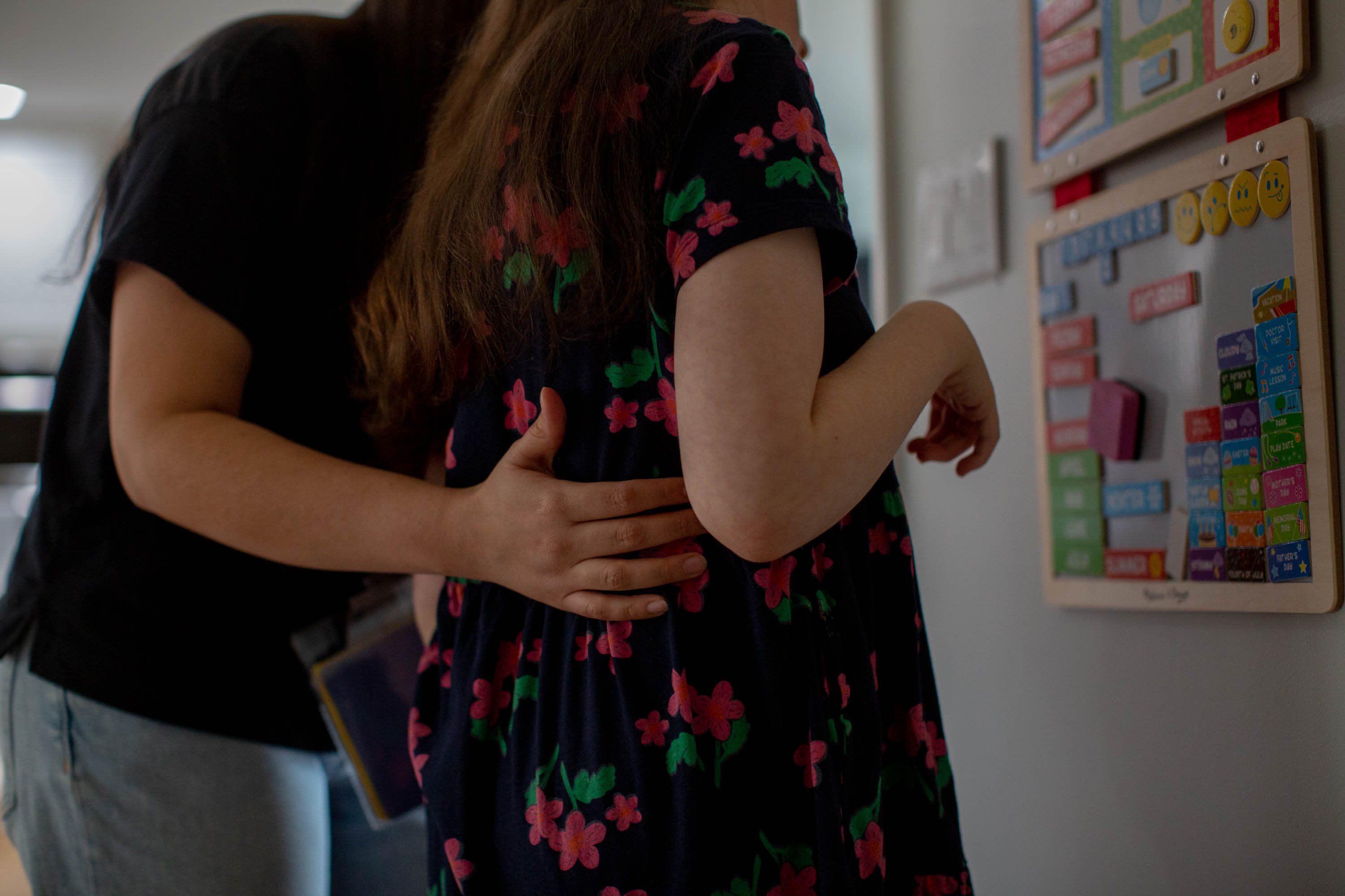 immigrant-caregivers-07 A woman in a black shirt holds her arm along the back of a girl wearing a flower dress as they look at a wall calendar together
