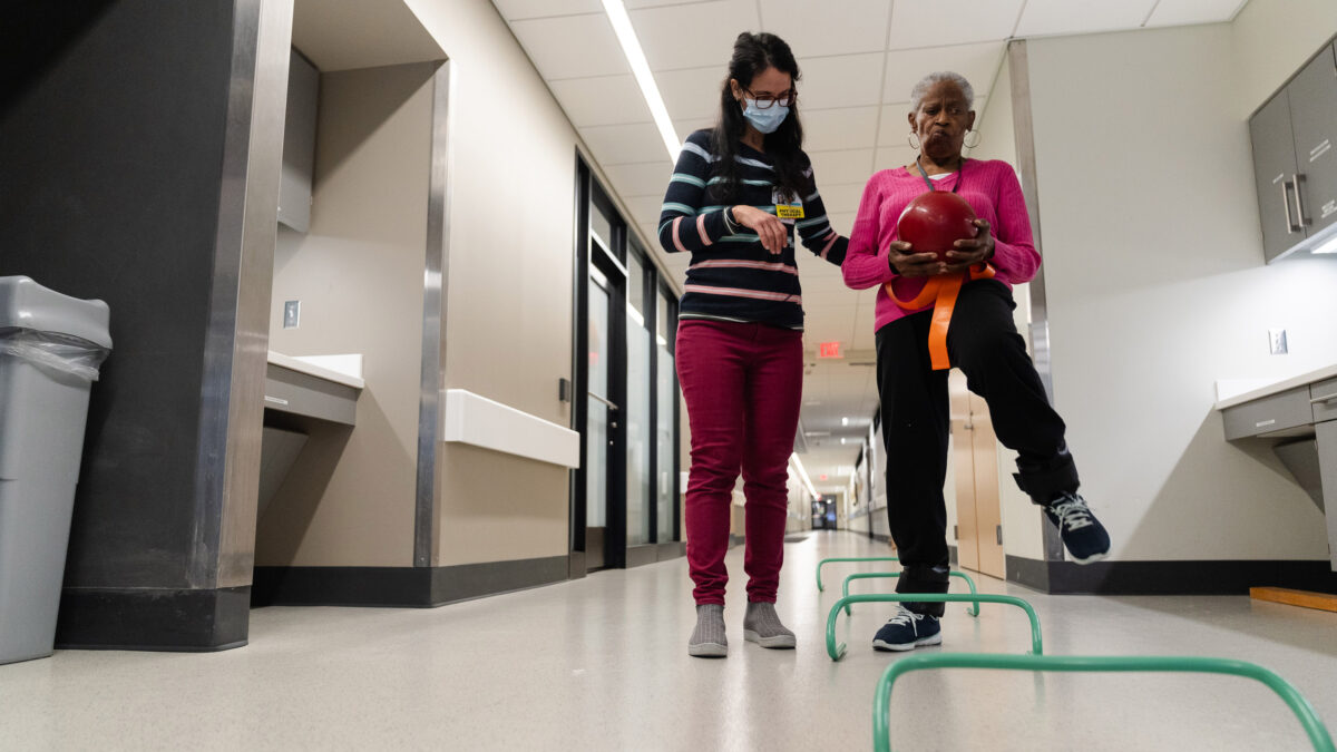A woman wearing glasses and a mask helps another woman balance on one leg while holding a ball