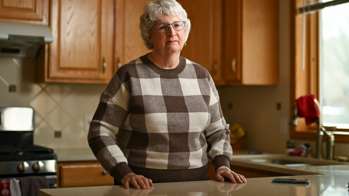 An older woman wearing glasses stands in her kitchen.