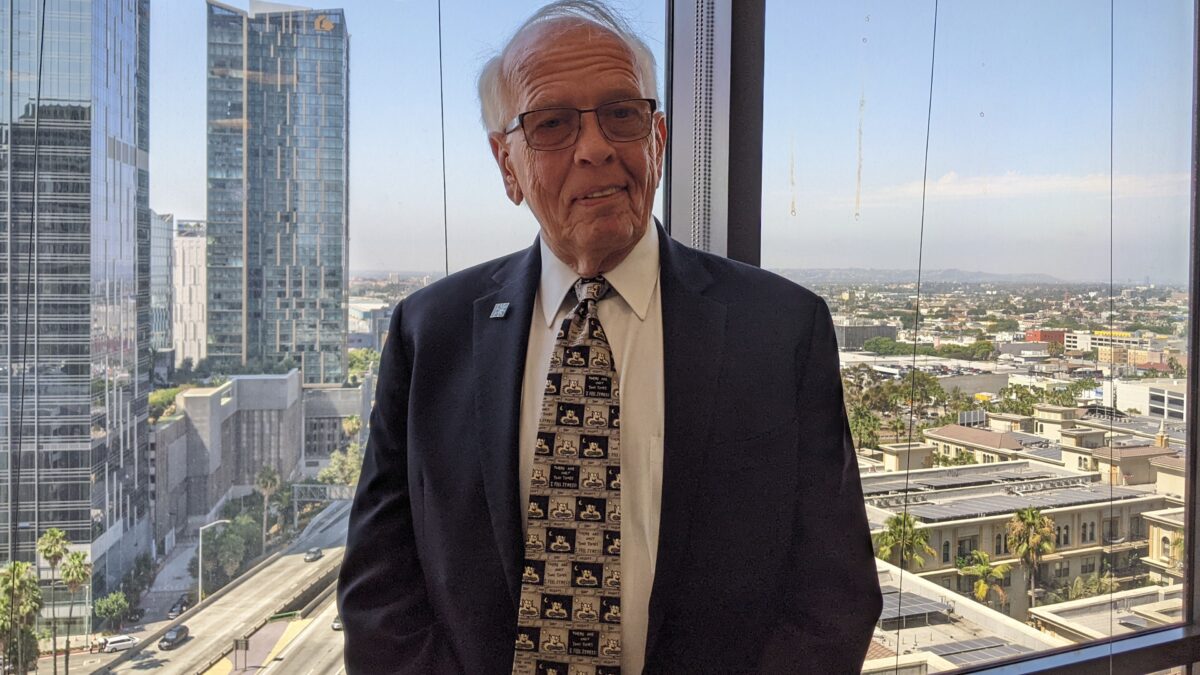 An older man in a suit stands in front of a large window with a view of skyscraper buildings