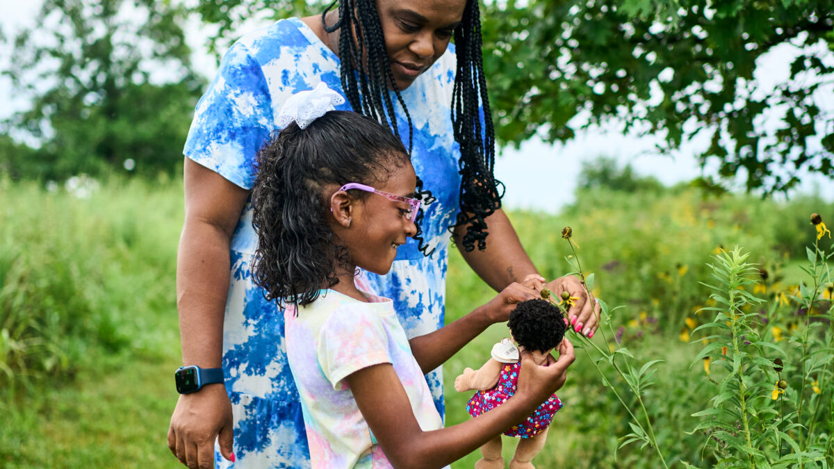 A photo of a mother with her daughter outside. The young girl is holding a doll and looking at flowers with her mom.