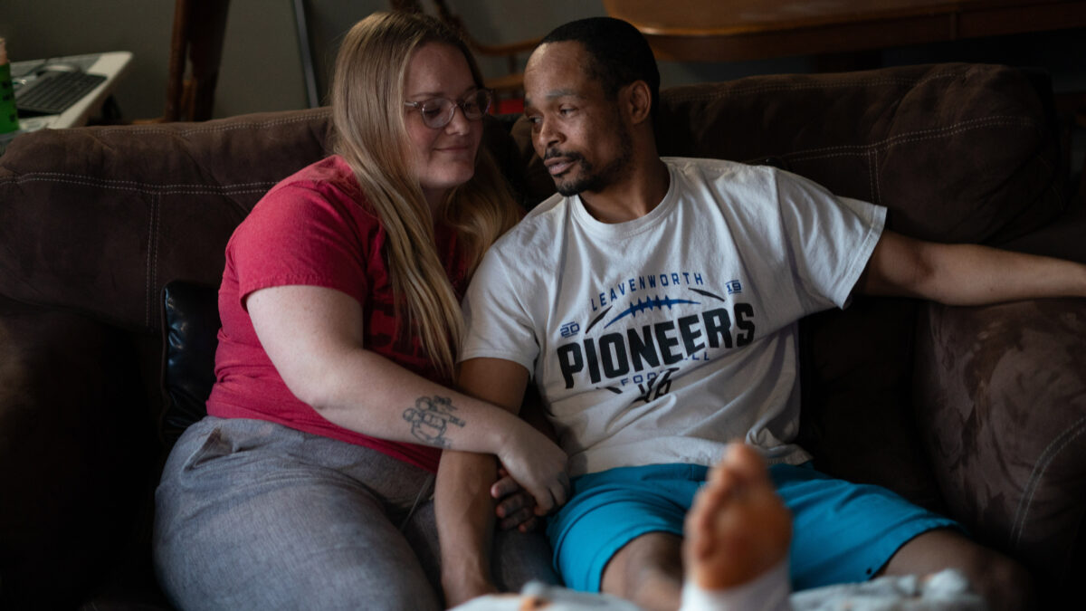 A woman and man sit on a couch, smiling at each other, with arms intertwined