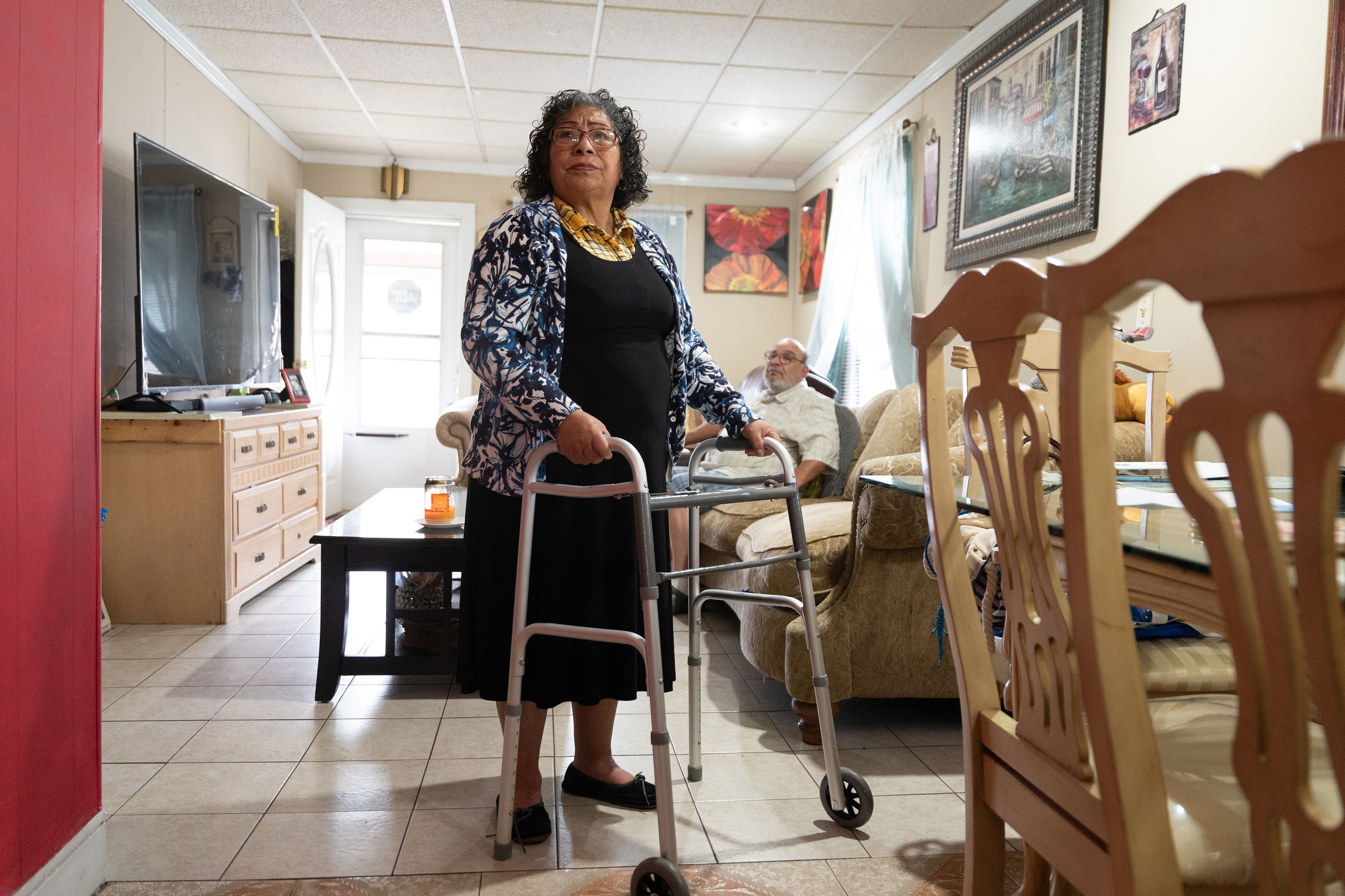 kansas-city-charity-money-06-3840 A woman wearing a black dress and blue and white shirt, holding on to a walker, stands next to a dining room table with a man sitting on a couch in the background