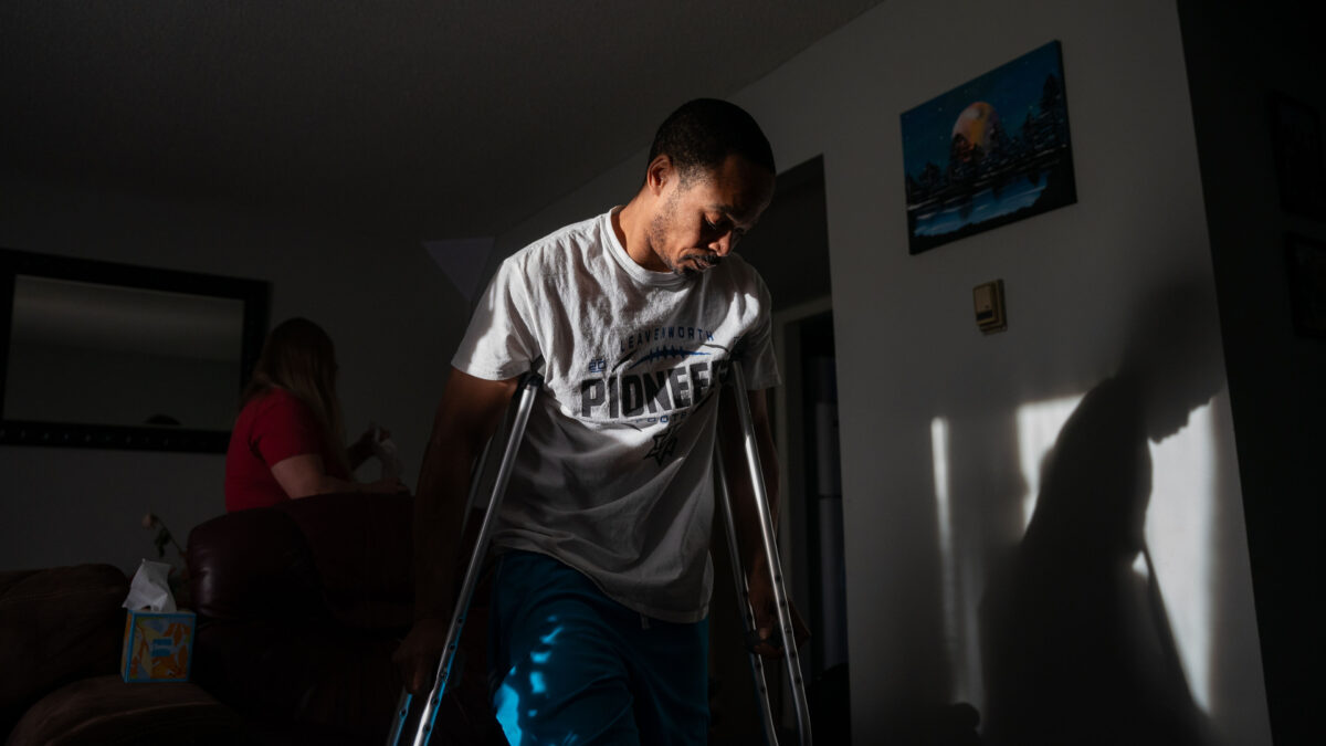 Jacob Gooch Sr. is standing on crutches in a room in his home. The room is dark, but he stands in a beam of light coming in through a window.