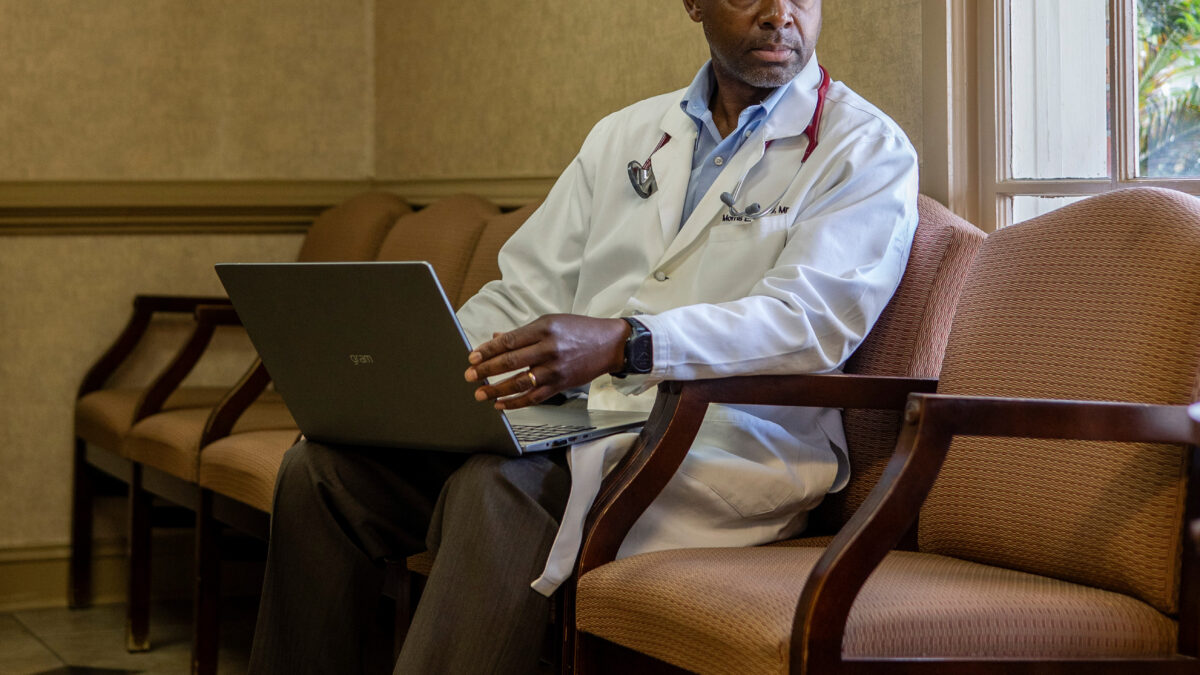 A photo of a Black doctor sitting in his office, looking to the right toward a window.