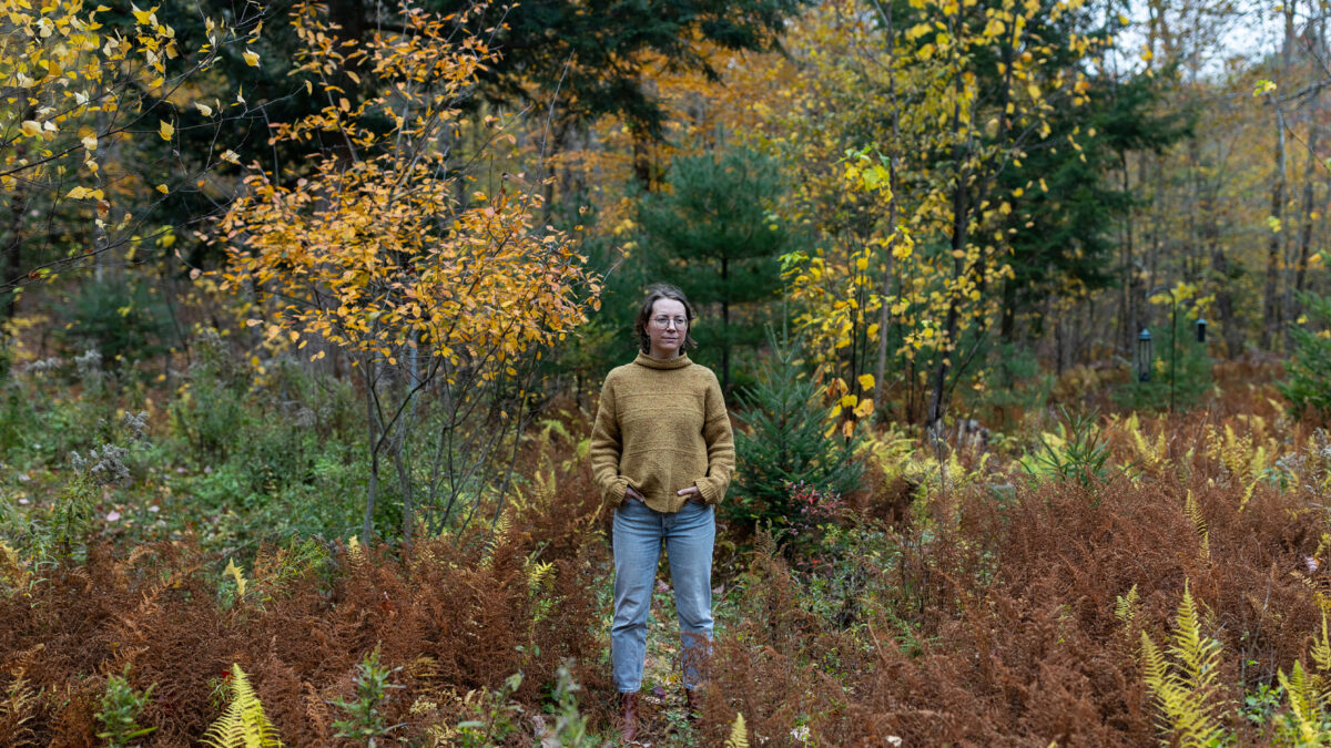 A photo of a woman standing in a meadow in autumn. The foliage around her is turning orange and yellow.