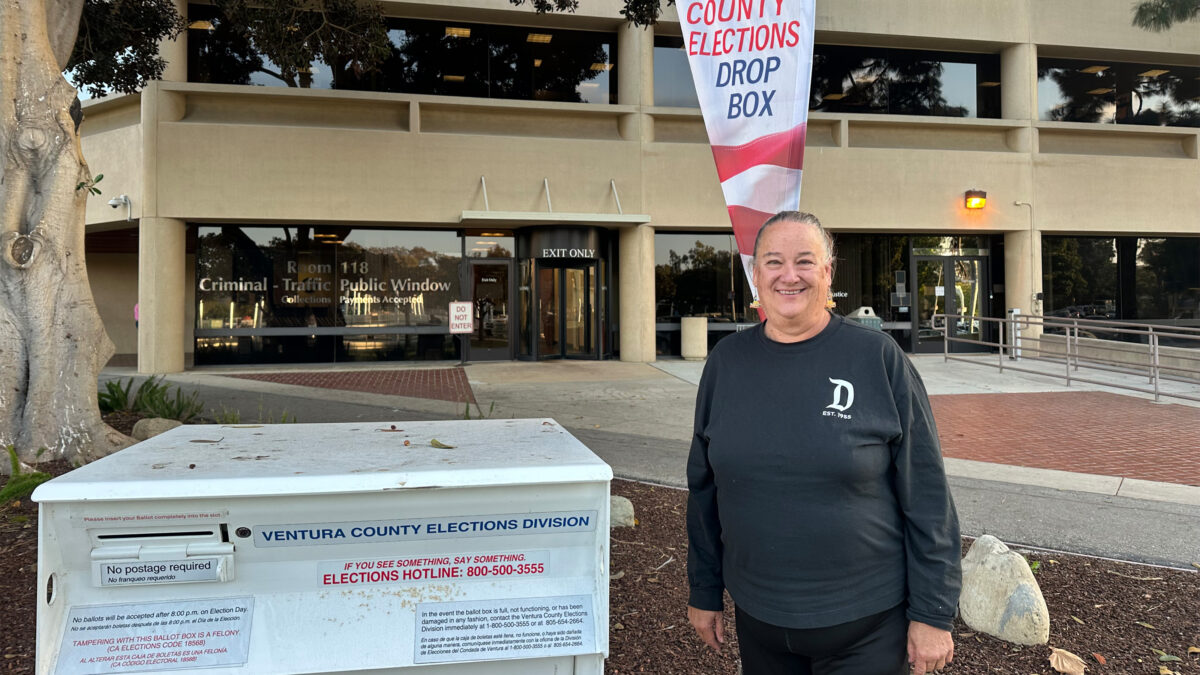A woman stands beside a ballot box, smiling.