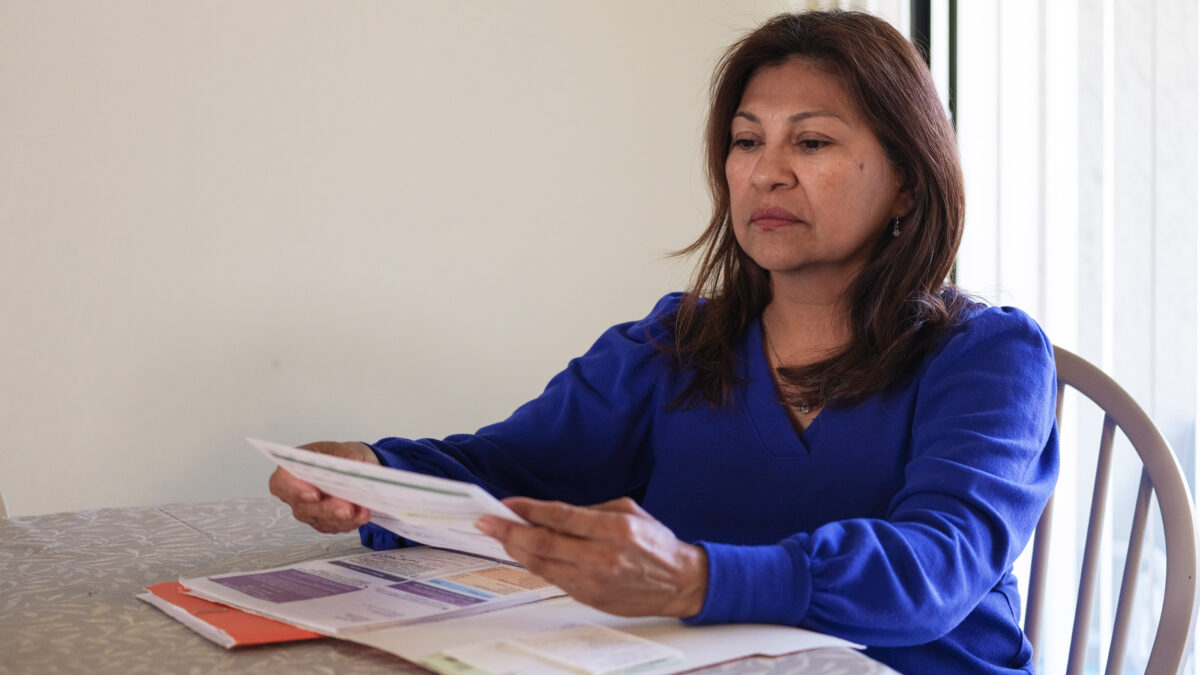 A Hispanic woman sits at a table, reading medical bills from a red file folder in front of her.